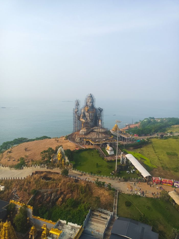 Aerial view of a grand Shiva statue surrounded by scaffolding, set against a serene ocean backdrop.